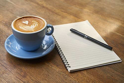 Eine Tasse mit Kaffee steht auf einem Holztisch, daneben liegt ein Notizzettel und ein Schreibtisch.