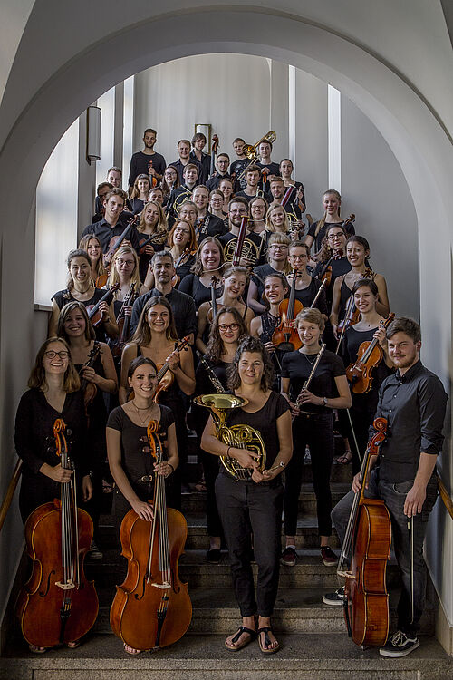 Auf einer Treppe stehen alle Mitglieder des Orchesters. Vorne die Streicher wie Celli und Geigen, hinten die Bläser wie Hörner und Trompeten.