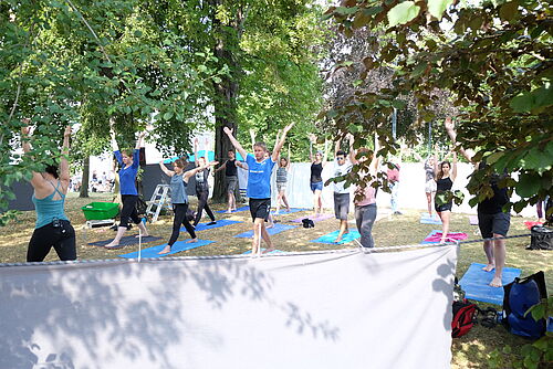 Im Bild befinden sich Teilnehmerinnnen und Teilnehmern im Stand mit nach oben gestreckten Armen in einer Yogastunde. Der Yogabereich wird im vorderen Bereich durch Tücher abgegrenzt.